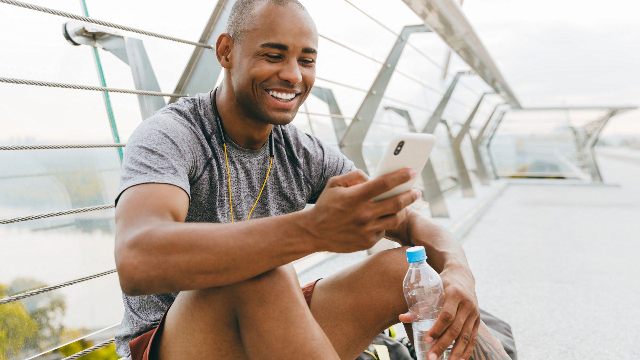 Man in sports gear looking at mobile on a cable bridge holding a bottle of water smiling